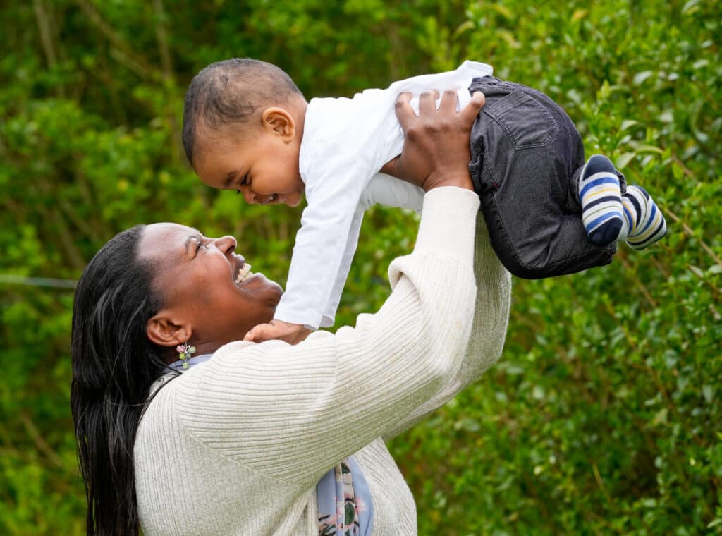 A smiling black mother lifts her one year old son into the air. He is laughing.