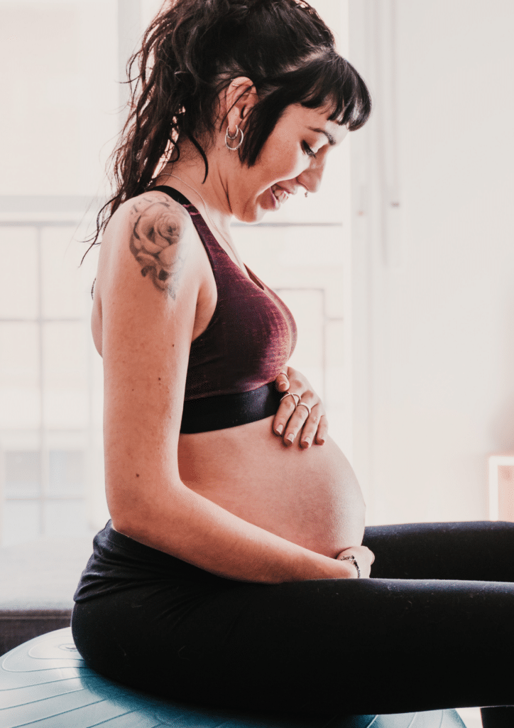 A pregnant woman with a tattoo on her arm is holding her belly while sitting on a birth ball