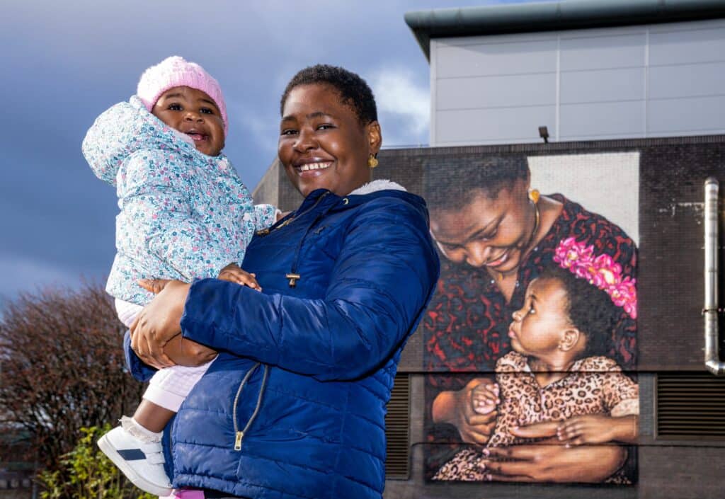 Black woman holding daughter and standing in front of a mural of her image